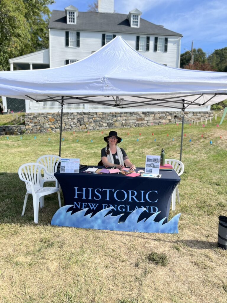 A woman sits under a white canopy at a "HISTORIC NEW ENGLAND" table in front of a historic house. Transcribed Text: HISTORIC NEW ENGLAND