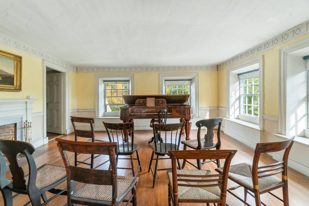 A room with a piano and wooden chairs, featuring yellow walls, large windows, and a fireplace.