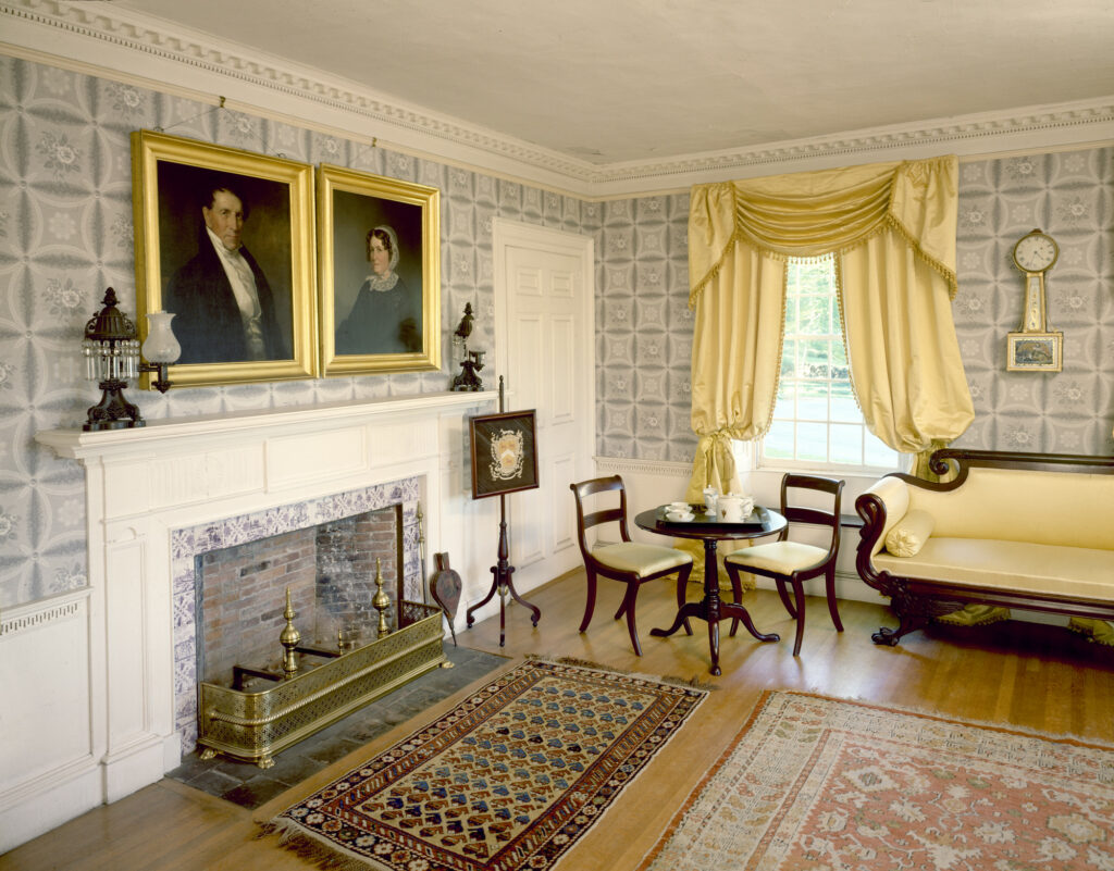 A classic 19th-century living room with a white fireplace, gold-framed portraits, patterned wallpaper, and antique furniture.