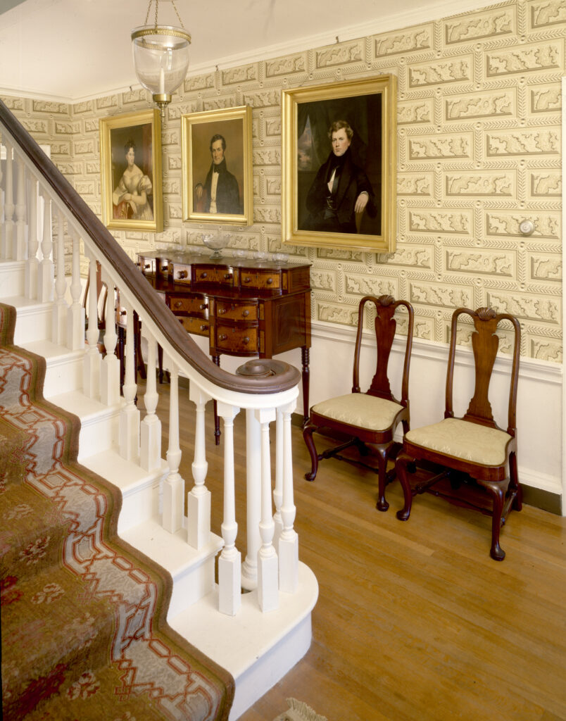 Interior hallway with a staircase, framed portraits, wooden sideboard, and chairs.