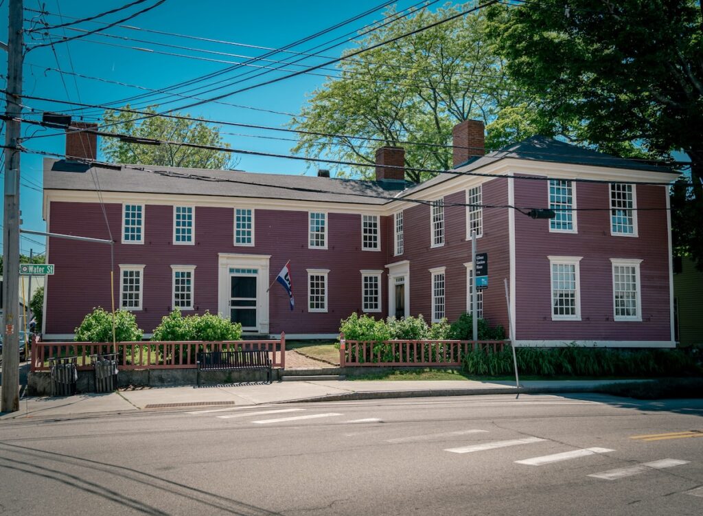 The image shows a two-story, dark red-painted colonial-style building with white window trim and two large brick chimneys. The building forms an L-shape and features a wooden fence in front with neatly trimmed greenery and plants.