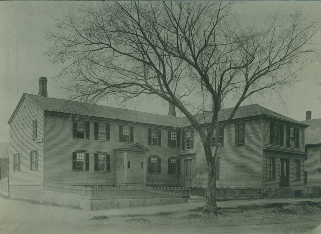 A large two-story wooden house with a gabled roof and a leafless tree in front.