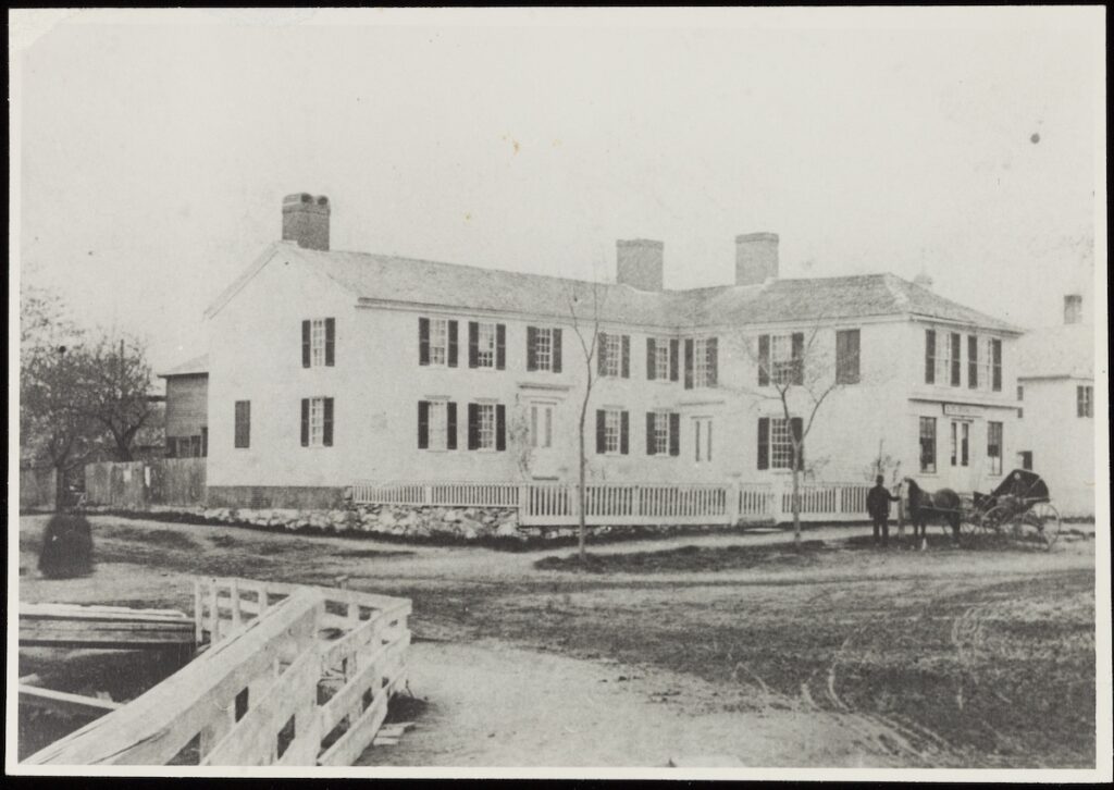 A two-story building with dark shutters and multiple chimneys, surrounded by a picket fence and dirt road with horse-drawn carriage.
