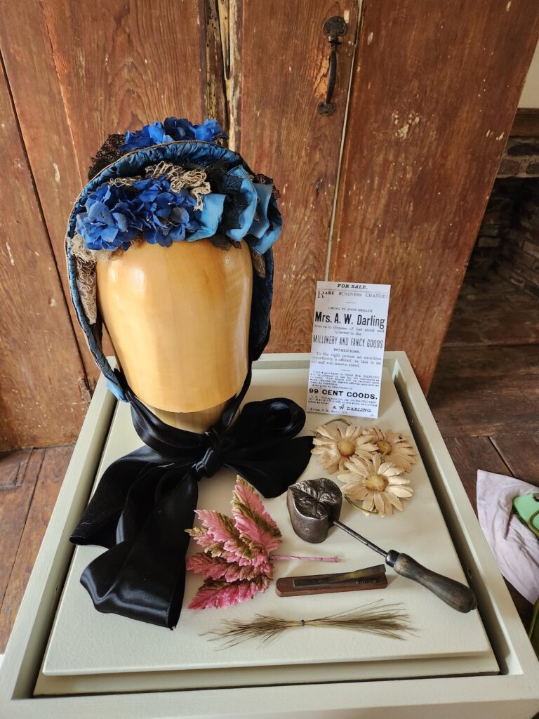 Mannequin head with a decorative headdress on a stand with vintage millinery tools and flowers.