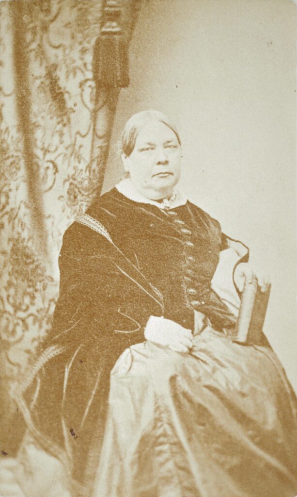 A sepia-toned portrait of a woman from the 19th century holding a book, seated with a patterned curtain in the background.