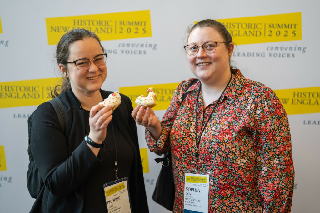 Two people smiling, holding sprinkle-covered pastries at the Historic New England Summit 2025 backdrop.
