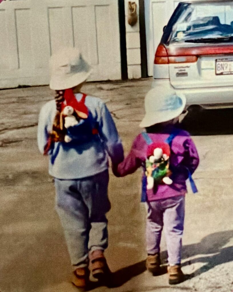 Two children in sun hats and backpacks hold hands, walking on a driveway next to a gray garage.