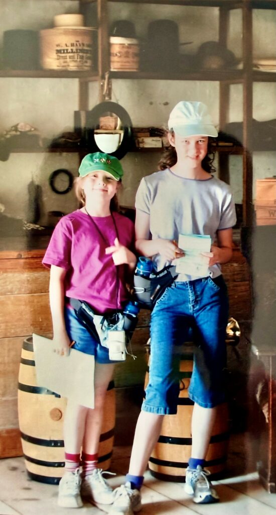 Two children in casual attire stand in front of wooden barrels, with shelves of vintage containers behind them.