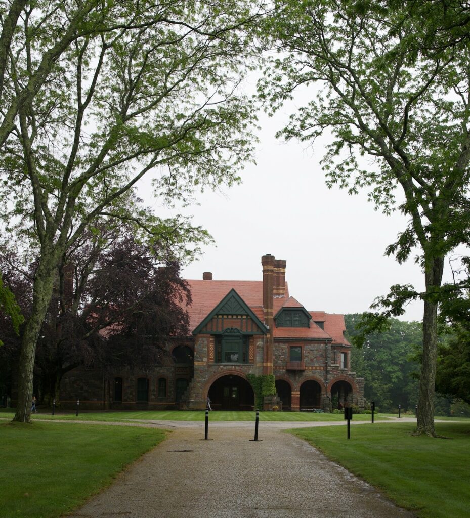 Historic stone mansion with arched entryways, a red roof, and lush greenery in the foreground on an overcast day.