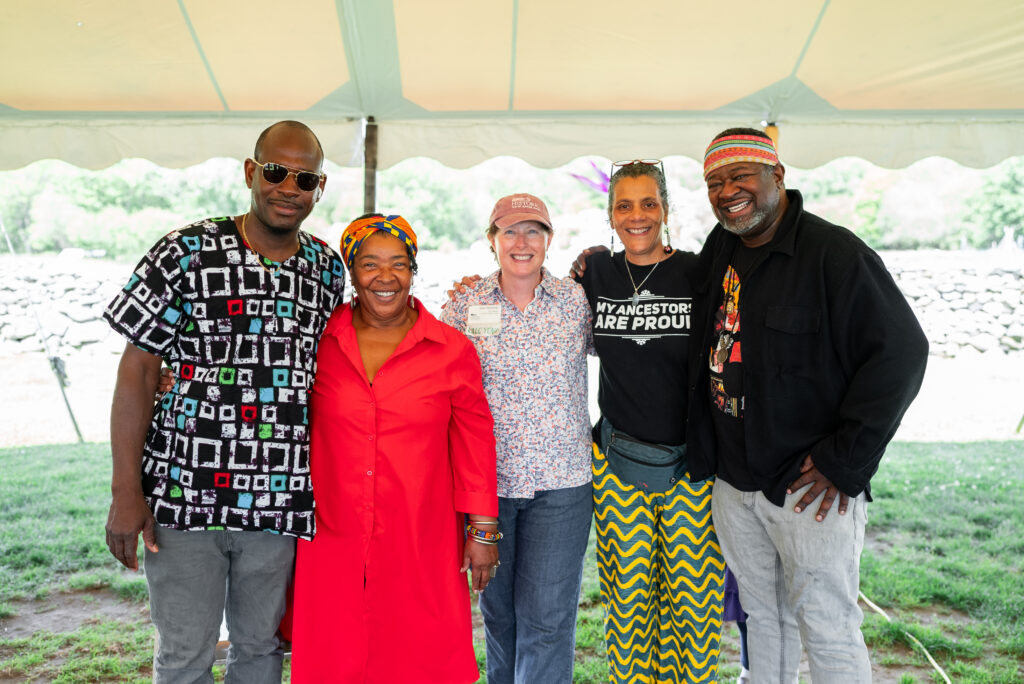 Group of five people smiling under a white tent, dressed in vibrant and patterned clothes. Lush vegetation in the background.