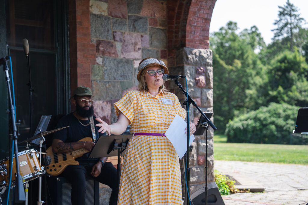 A person speaks into a microphone outside a historic stone building. A musician sits behind, surrounded by green trees.