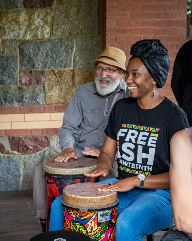 Two people happily drumming together on a porch with rustic brick and stone walls in the background.