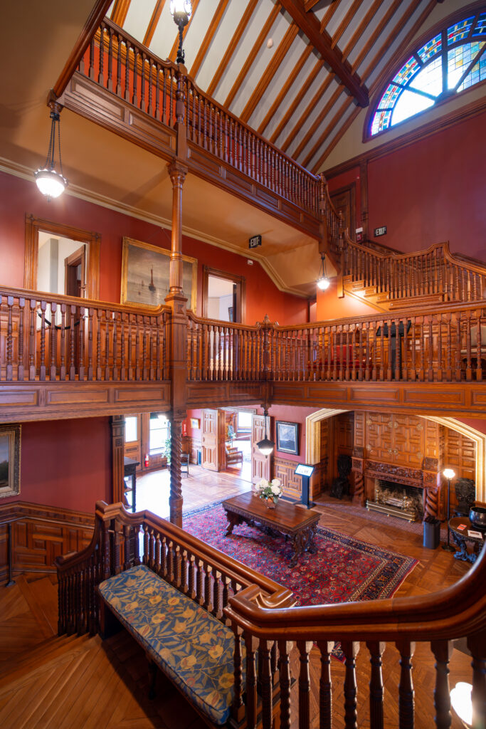 Grand wooden staircase and balcony with ornate detailing, rich red walls, and a stained glass window above.
