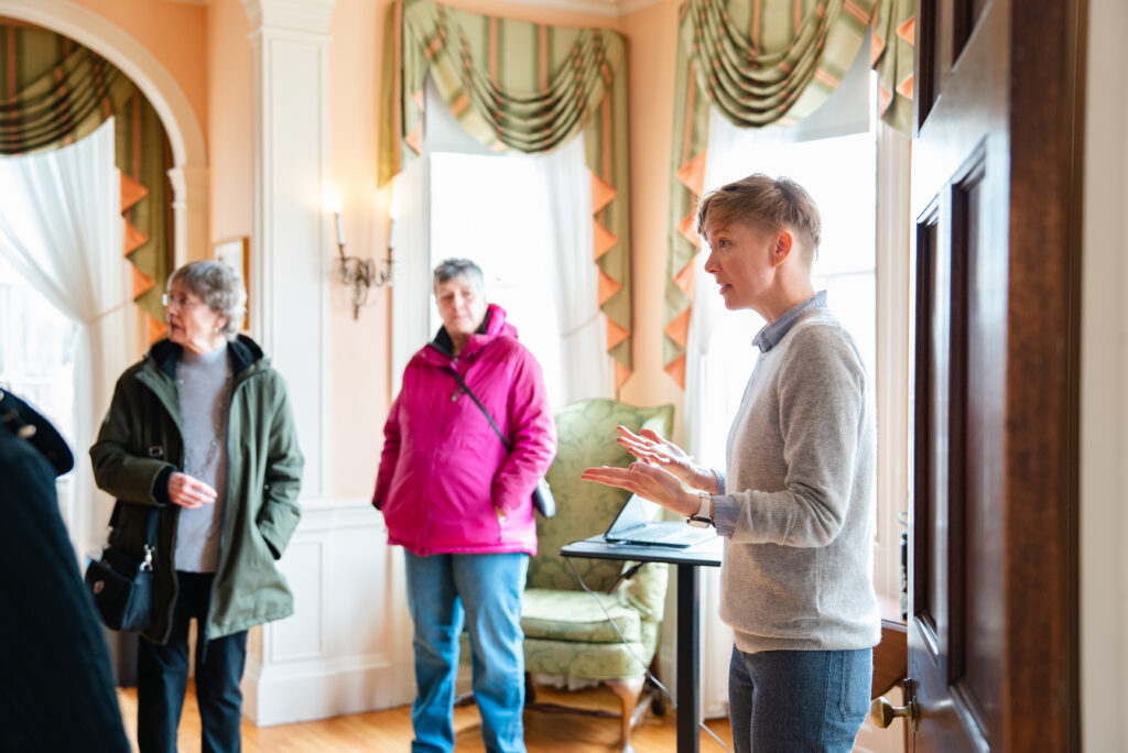 Guide gesturing in a historic room with decorative drapes, sconce lighting, and visitors observing.