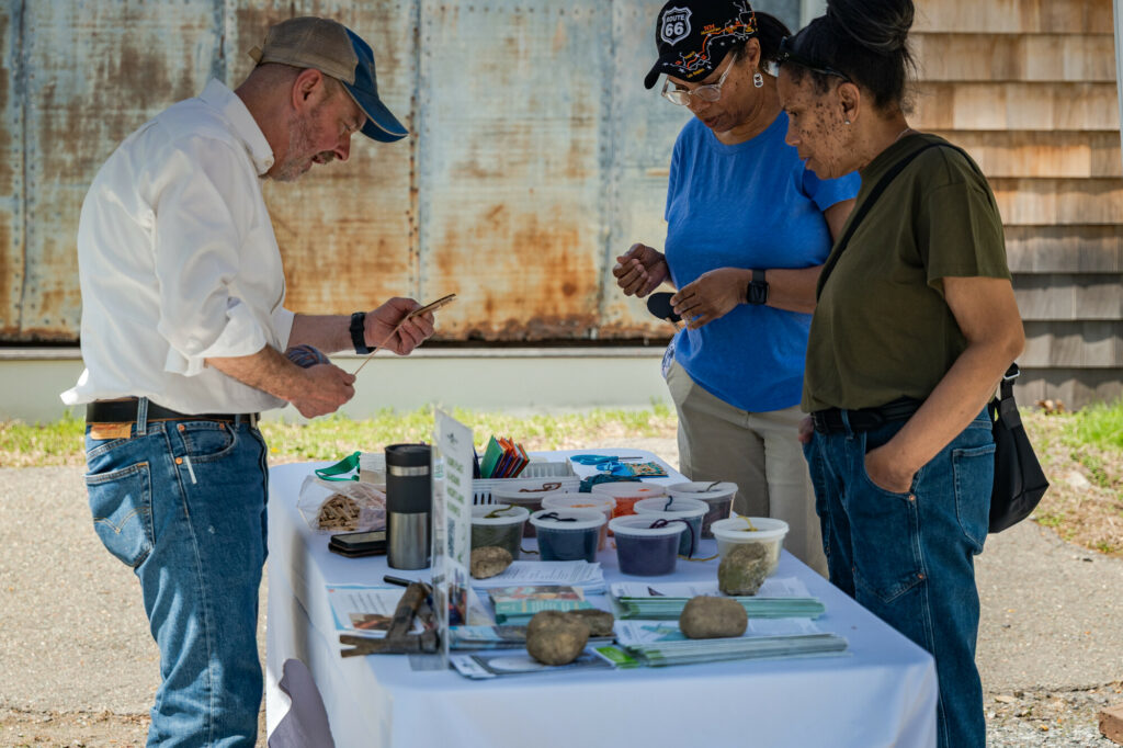 Outdoor table with art supplies and plants; three people engaging in conversation and examining items.