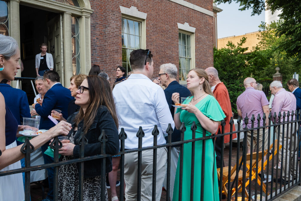 People mingle at an outdoor gathering by a historic brick building with large windows and lush greenery nearby.