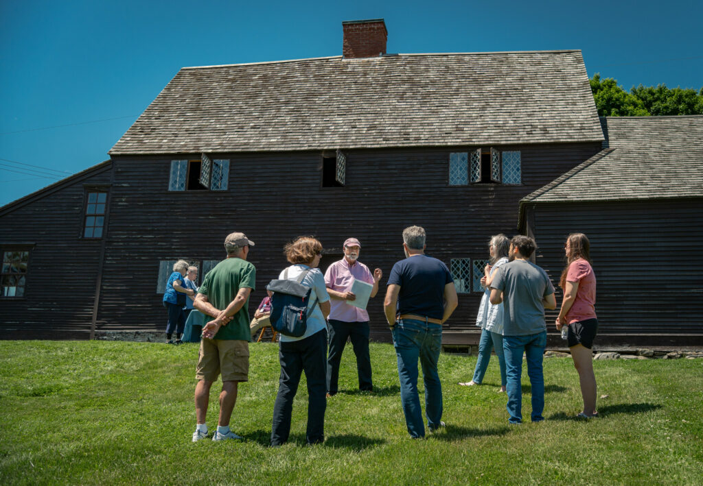 A group tours a historic, dark wood colonial house with diamond-pane windows, standing on a lush green lawn under clear skies.