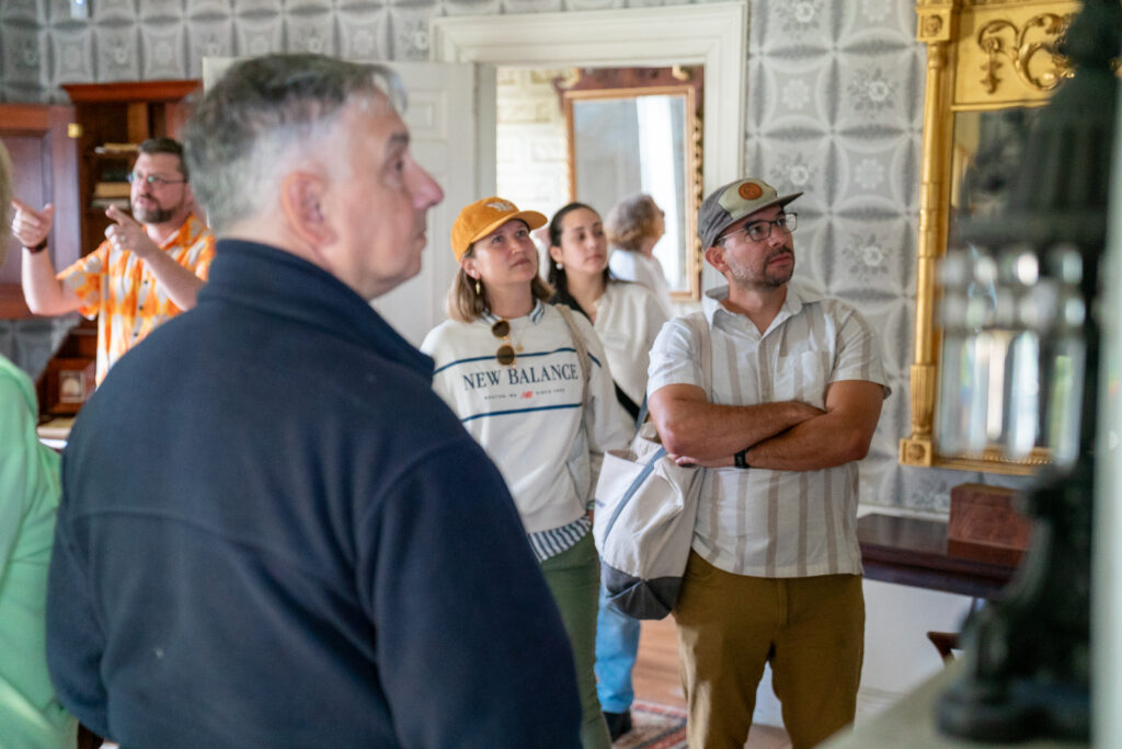 People tour a room with ornate wallpaper, a carved mirror, and a wooden bookshelf, as a guide gestures to features.