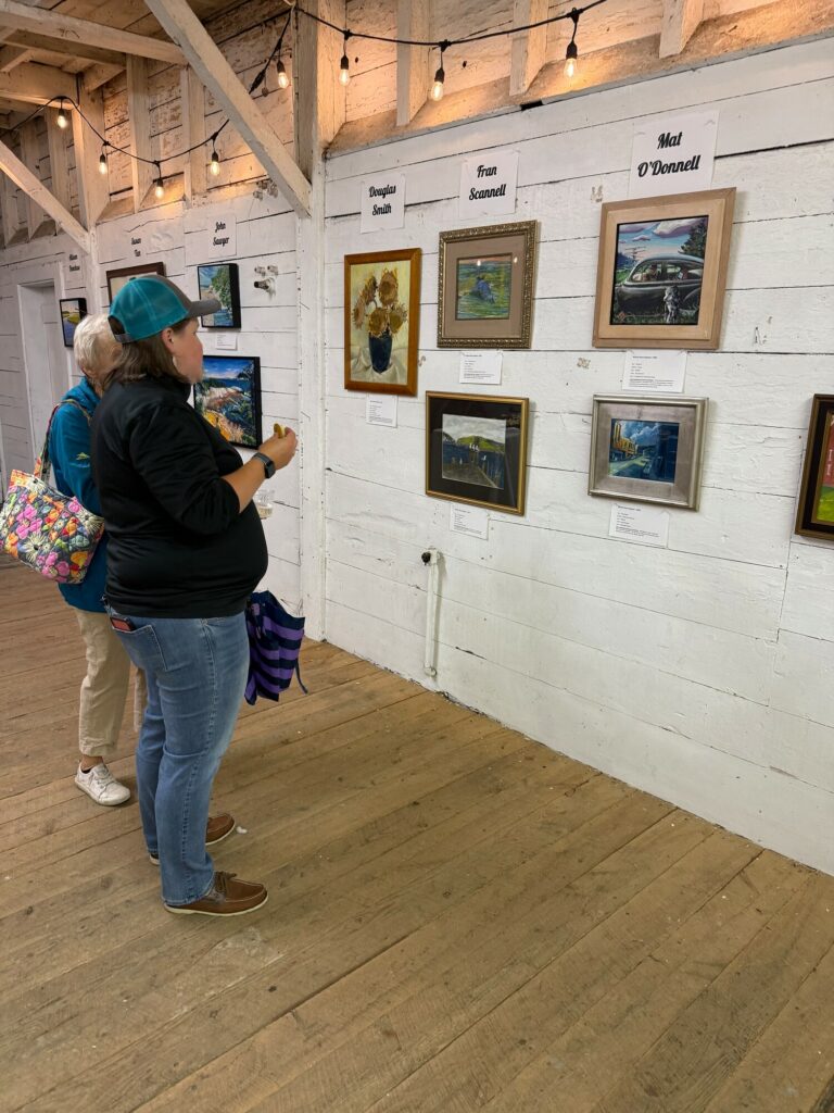 Two women observe framed paintings on a white wooden wall, beneath string lights in a rustic gallery space.