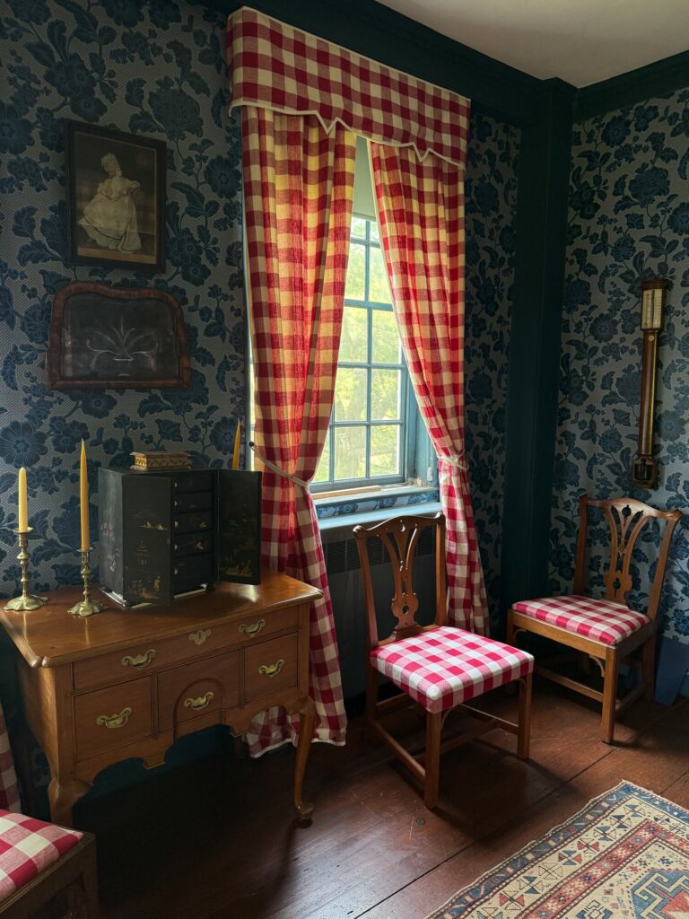 Colonial-style room with checkered curtains, blue floral wallpaper, wood furniture, and a cozy bed with a canopy.