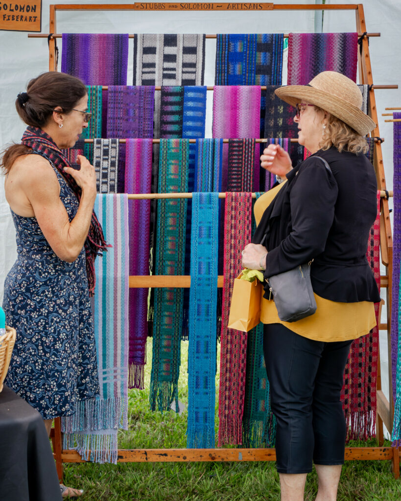 Two women admire vibrant, woven scarves displayed on a wooden rack at an arts and crafts fair.