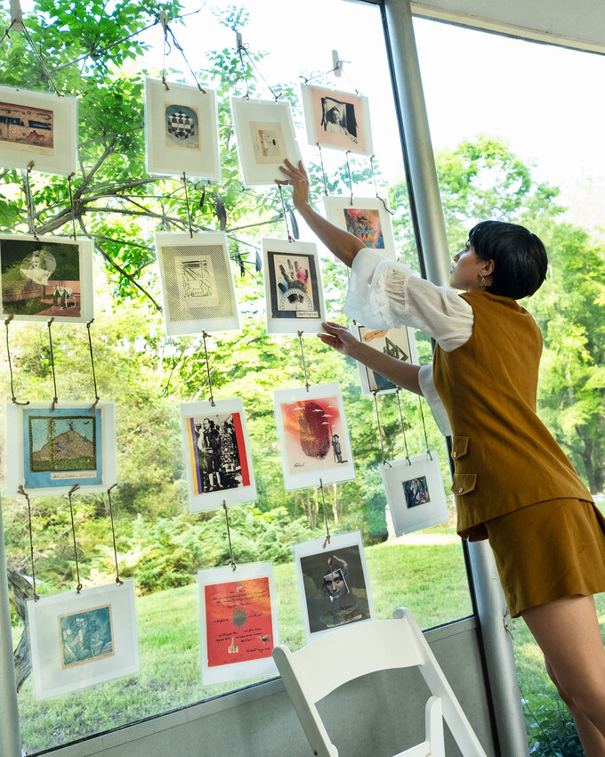 A person arranges framed art on a window, with a garden visible outside. A white chair sits in the foreground.