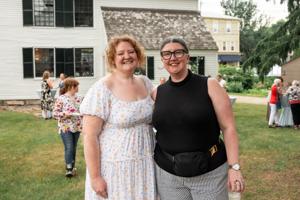 Two people pose happily in front of a white colonial-style house, with lush greenery in the background.