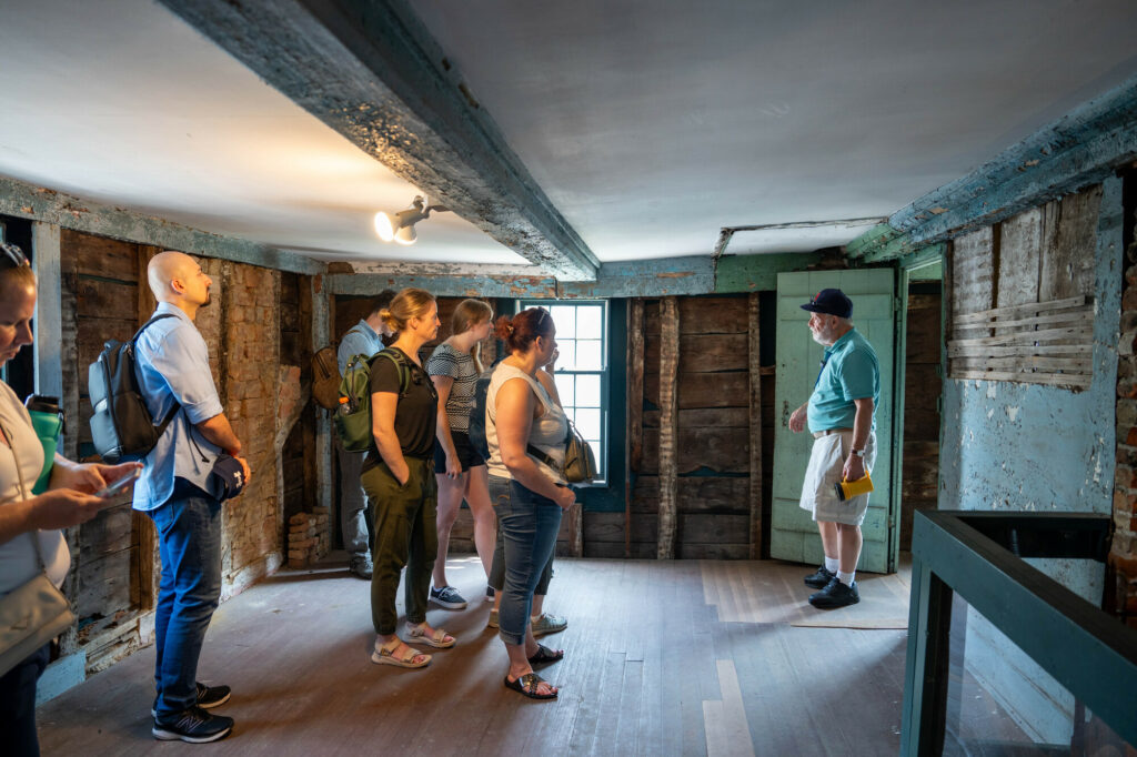 A group tours a rustic attic with exposed beams and brick, led by a guide in historical attire under soft lighting.