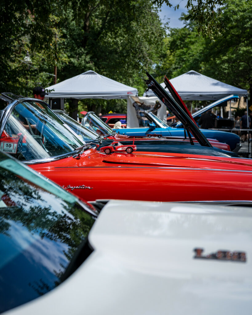Classic cars showcased outdoors; vintage red cars with model cars on hoods, surrounded by trees and tents.
