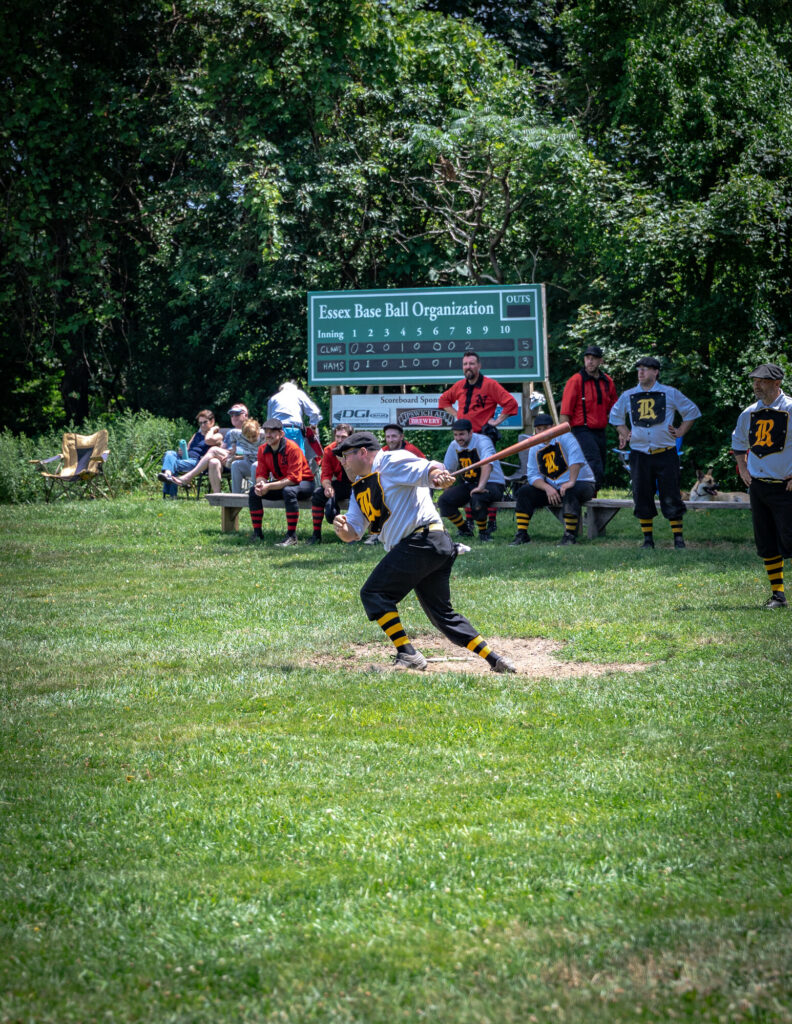 Vintage baseball game in progress; player swinging bat on a lush field, with scoreboard and spectators in background.