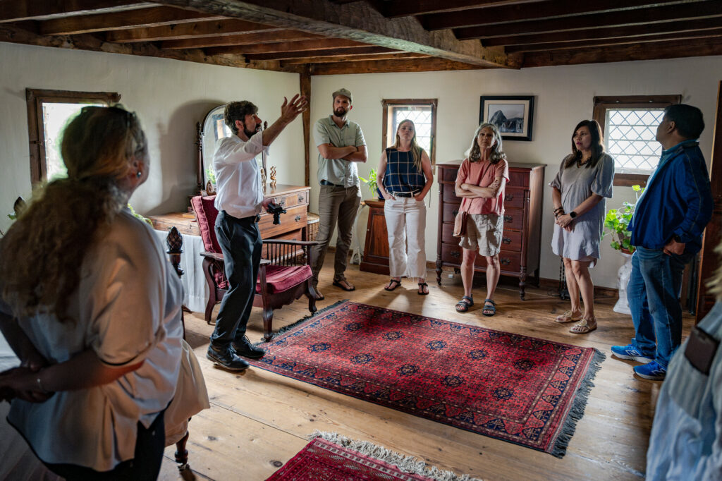A group of people in a rustic wooden room, with red oriental rugs and vintage furniture, engaged in a guided tour.