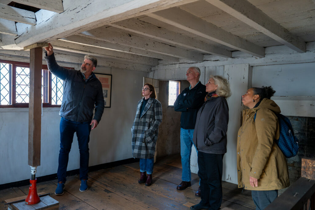 A guide explains wooden beams to a group in a historic room with checkered windows and rustic timber floors.