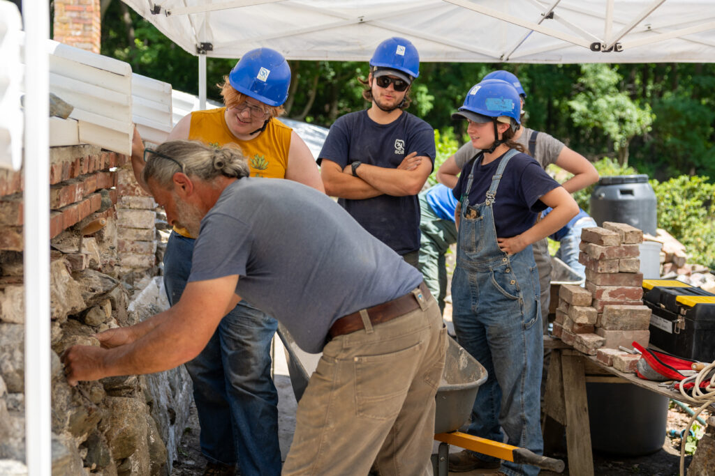 People in blue hard hats observe as a craftsman restores a historic brick wall under a white canopy. Lush greenery in background.