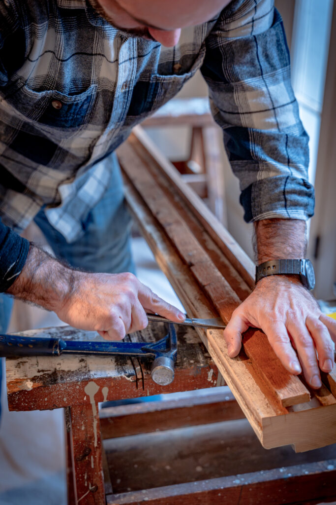 Artisan in flannel shirt chiseling wood on workbench, with blue-handled hammer nearby, crafting detailed architectural piece.