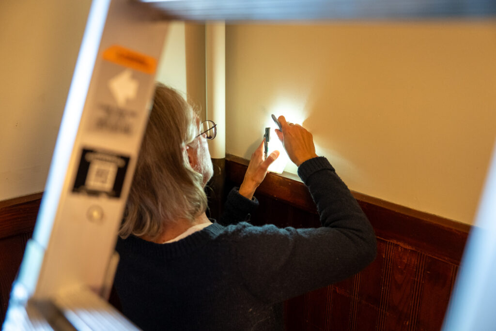A person adjusts a light switch near a wooden-paneled wall, viewed through a ladder.