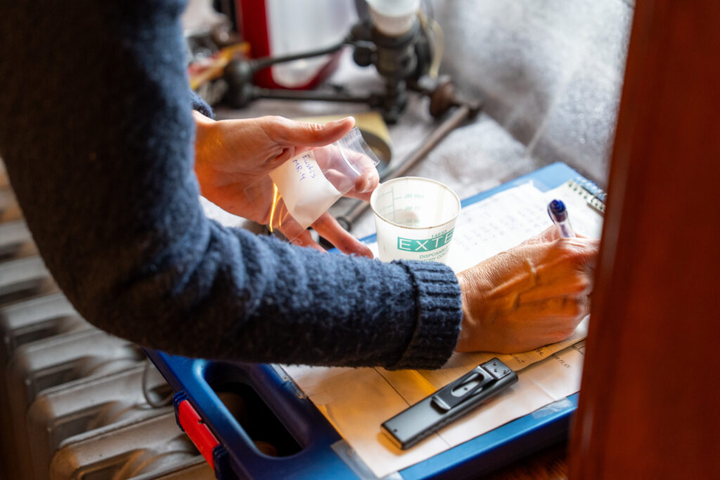 Hands handle a sample bag and cup, writing notes on a blue clipboard, with laboratory tools nearby.