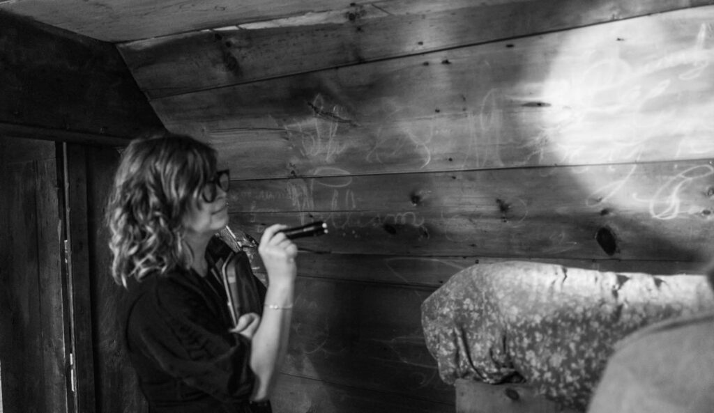 Woman with flashlight examines rustic wooden wall with faint chalk writings. Cozy fabric-covered bench in foreground.