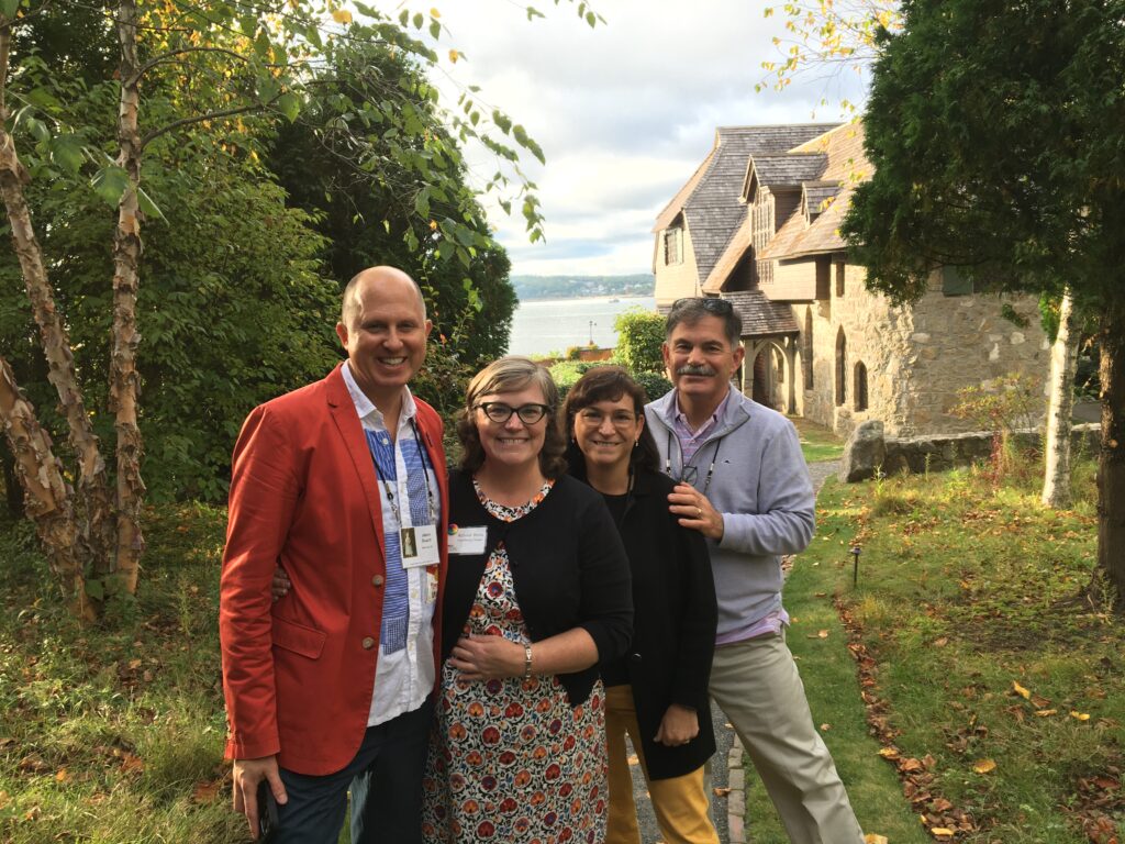 Four people stand smiling in a garden with lush greenery, beside a stone building overlooking a scenic water view.