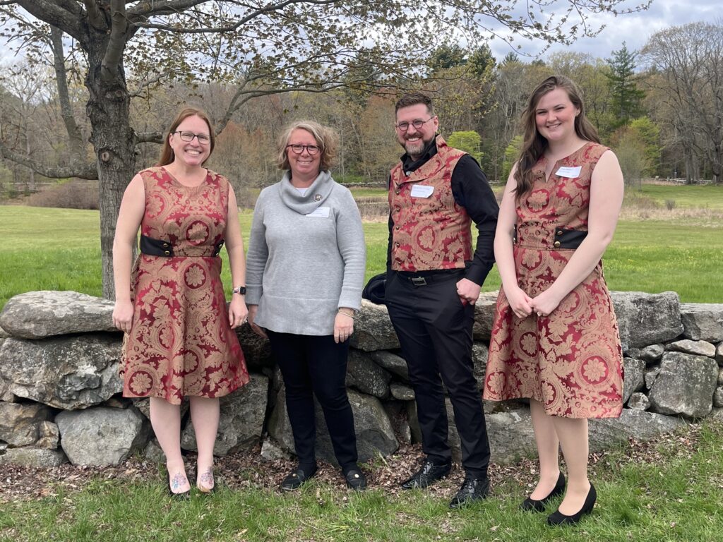 A group of people, three in matching red floral outfits, smile in front of a stone wall and lush green landscape.
