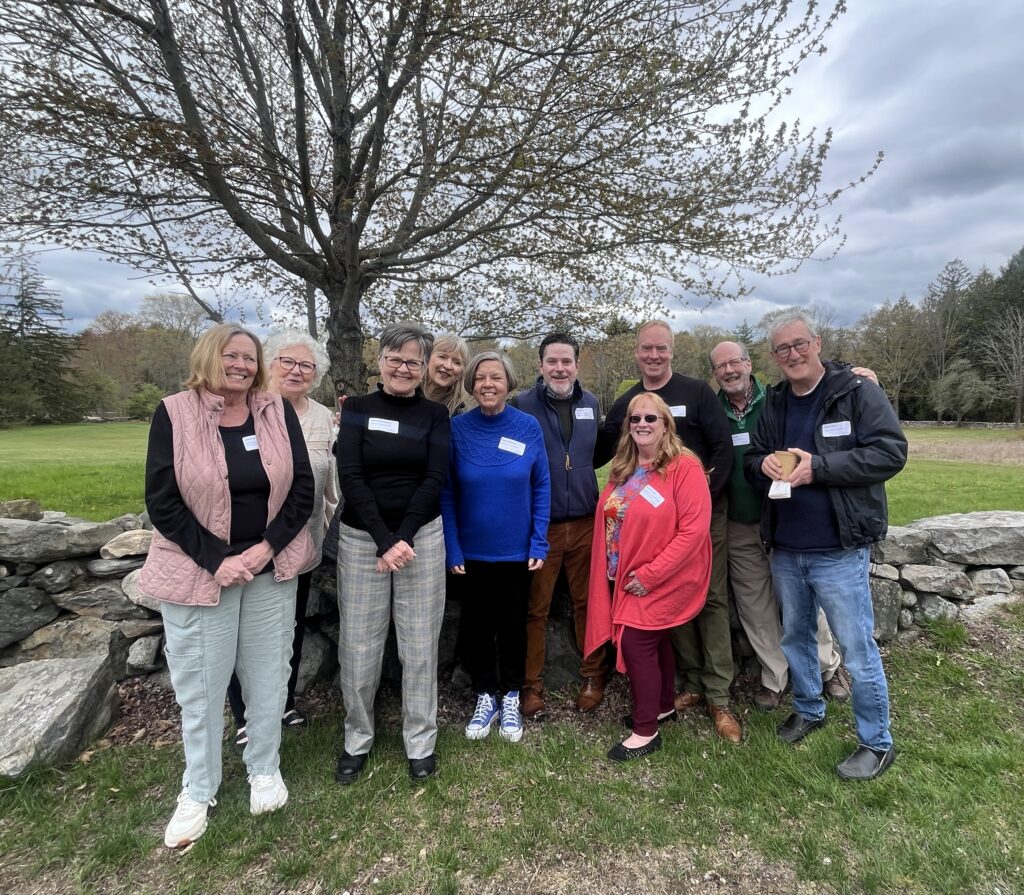 A group of smiling people stands in front of a stone wall and a tree in a lush, green park setting.
