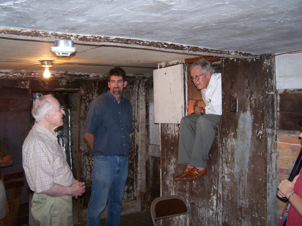 Three men in a rustic, historical basement; one peers out from a wooden hatch, with a low ceiling and weathered walls.