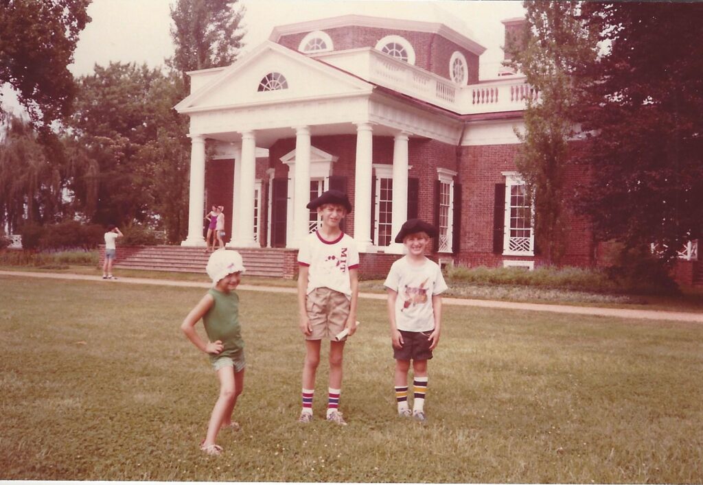 Children pose in front of a neoclassical red brick building with white columns and lush greenery surrounding them.