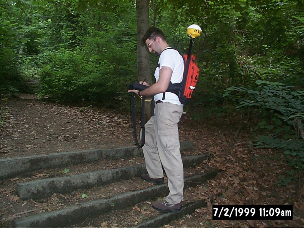 A person stands on forest steps, using equipment. Surrounding them are dense trees and leaves. Date: July 2, 1999, 11:09am.