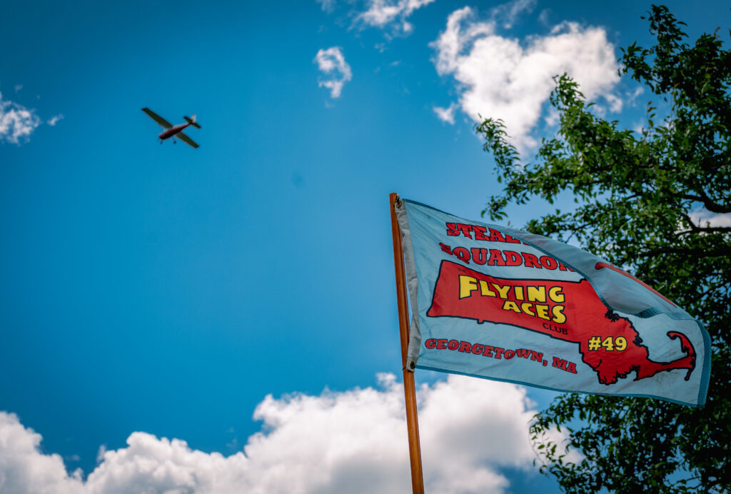 A small plane flies over a Flying Aces club flag against a clear sky, with green tree branches and fluffy clouds.