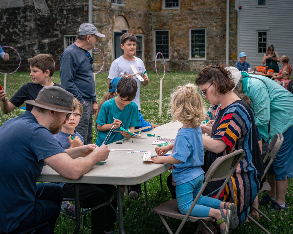 Families engage in outdoor crafts at a table, with a historic stone building in the background.