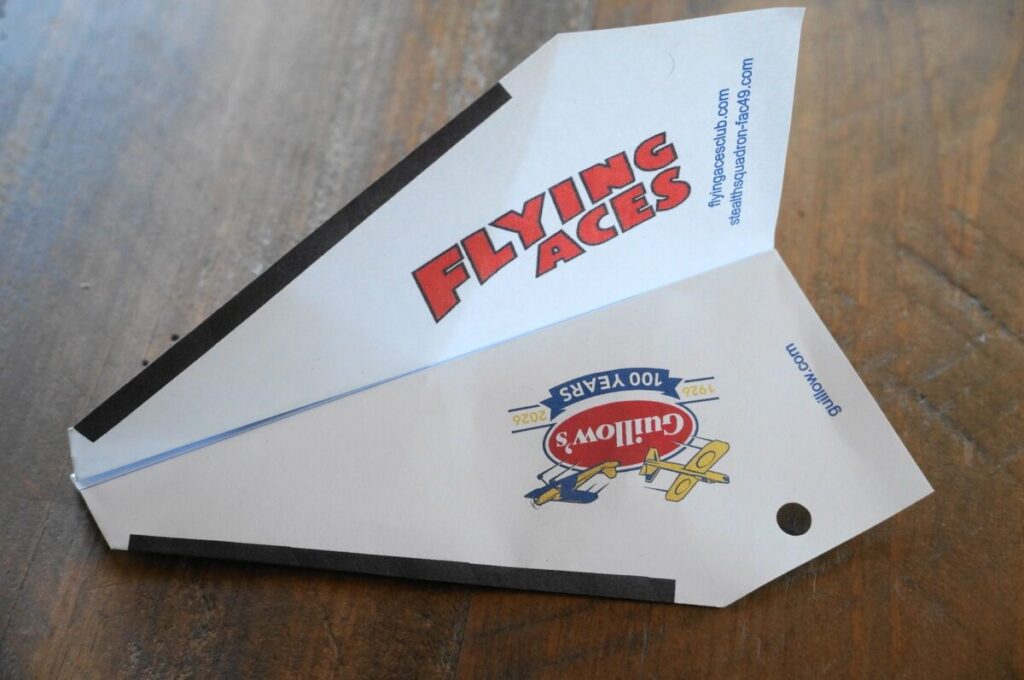 Paper airplane with Flying Aces text. Designed on a wooden table, it also features a logo with a vintage biplane.