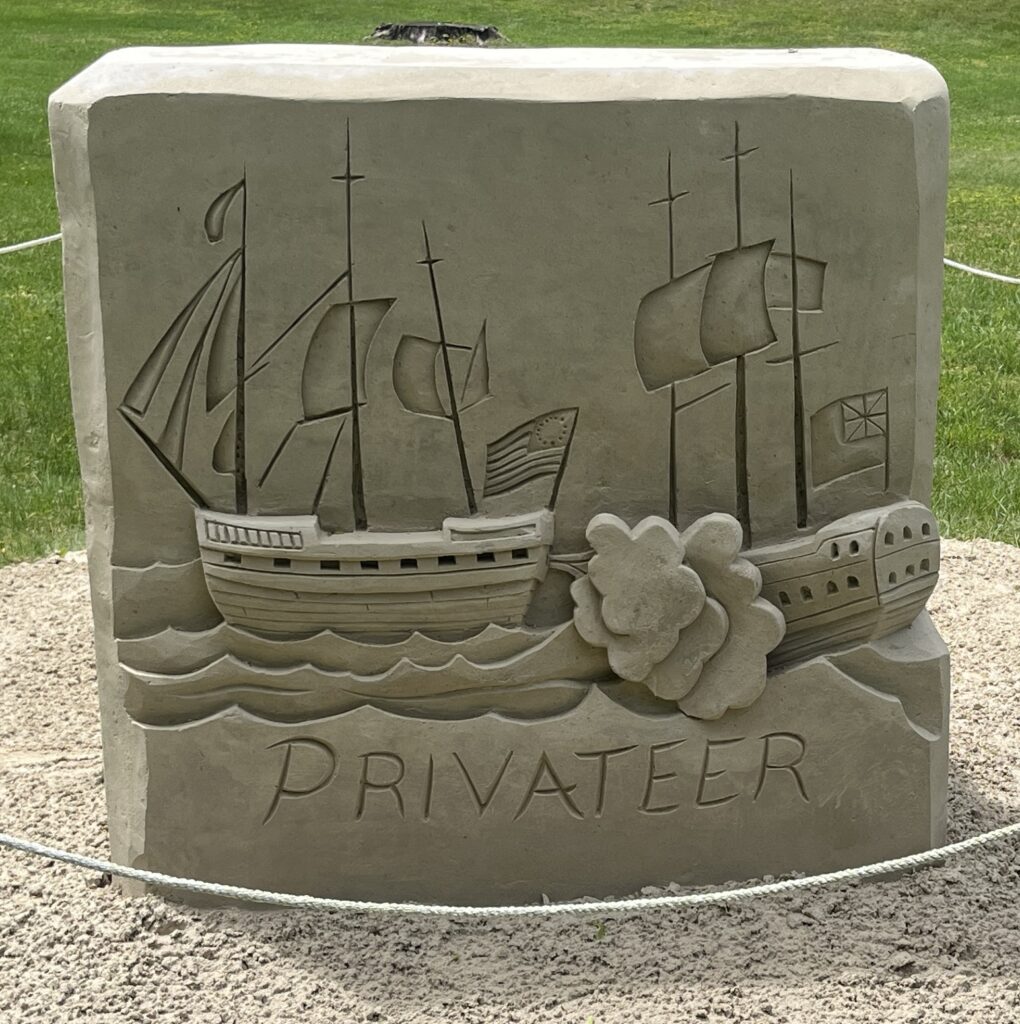 Sand sculpture depicting two sailing ships with waves and smoke, titled Privateer, set against a lush grass backdrop.