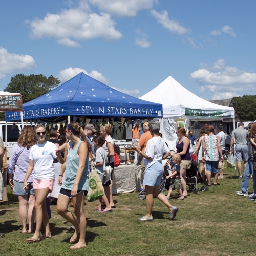 Crowded outdoor market with blue and white tents on a sunny day. People gather near Seven Stars Bakery tent.