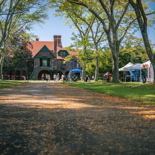 Historic manor surrounded by trees and visitors, showcasing eclectic architecture with red roofing and arched entry.
