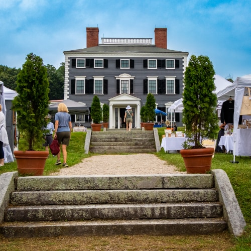 Historic three-story mansion with lush gardens, flanked by market tents. Stone stairs lead to the front entrance.